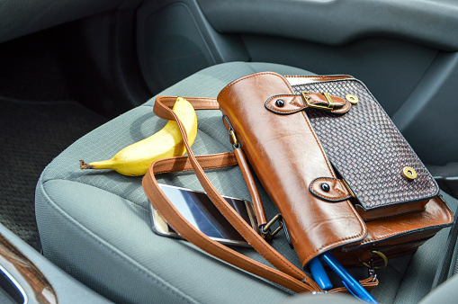 This is an image of a brown purse on the passenger seat of a car with contents spilling out.  There is a pen, makeup item, phone and banana laid out around the purse.  The car interior is grey.