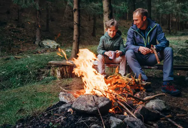 Photo of Father with son warm near campfire, drink tea and have conversation