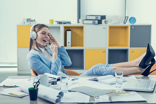 Young women with feet up on desk listening music on headphones and singing at office