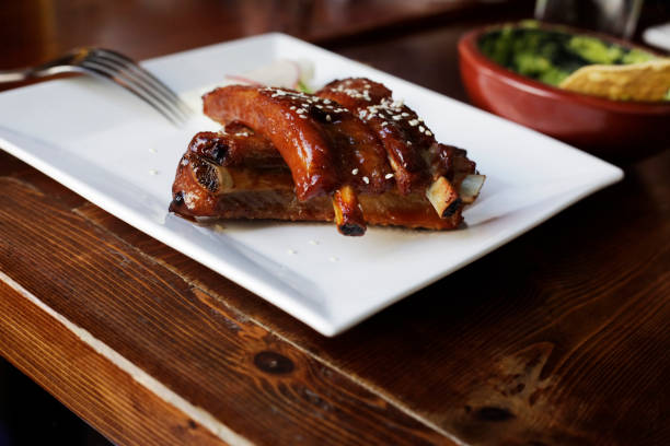 A plate of barbecued ribs, glazed in a rich sauce and marked with grill lines. The ribs are accompanied by a serving of golden French fries, a piece of corn on the cob with a toothpick in it for easy handling, and a small portion of purple coleslaw on a bed of lettuce.