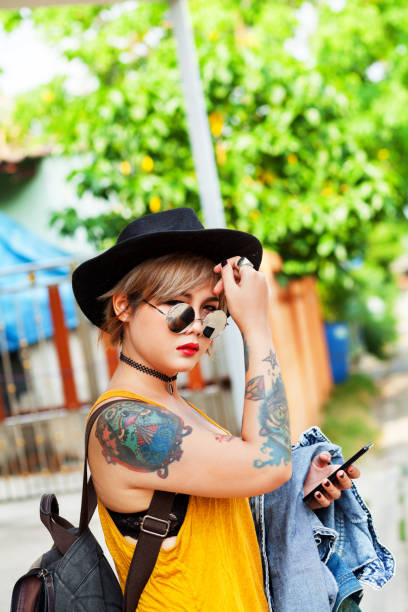 Thai girl ready for travel: thai girl standing with backpack and mobile wearing hat in street. Girl has tattoos. Street scene in Bangkok.