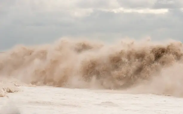 Giant wave breaking on a beach. Water appears to be muddy. Lots of fine spray (is not noise). Sea foam on the left. Overcast sky. Lots of detail.