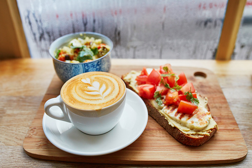 Delicious breakfast, brunch or lunch with hummus and tomato sandwich, salad and fresh hot cappuccino coffee served on cutting board near the window with water drops on wet glass at rainy day