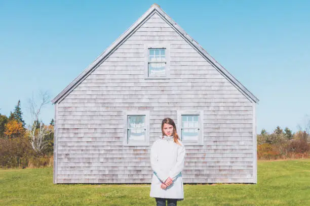Young girl in white trenchcoat standing with in front of her home, looking towards the camera. Abstract Concept Portrait. Nova Scotia, Canada.