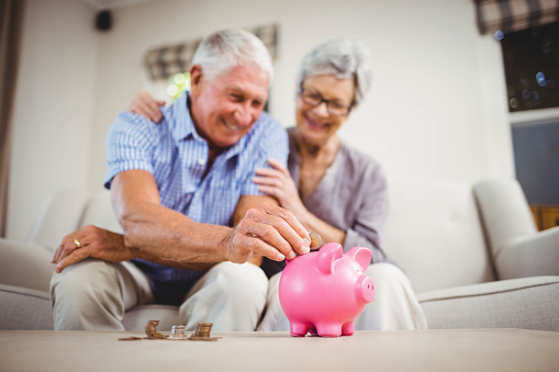 Senior man sitting with woman on sofa and putting coins in piggy bank