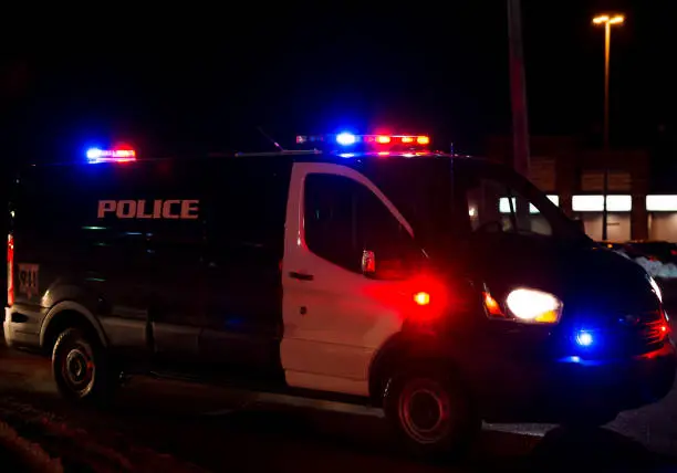Police van at night. Lights on it are flashing. Van is black and white, door is very dirty. Road is dirty and has some slush. Focus on light bar. Van is generic, brand marking have been removed. Cannot identify model.