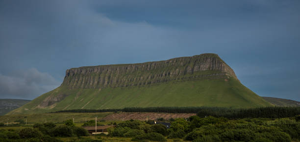 A strange table-shaped mountain in ireland's rural landscape In County Sligo we find a table-shaped landscape: Benbulben Mountain. This giant Ice Age massif is easily recognizable by its atypical shape: its steep limestone slopes are almost perpendicular to the ground with large cracks that look like ribs. A soft green grass covers the mountain giving it an even more beautiful look. ben bulben stock pictures, royalty-free photos & images
