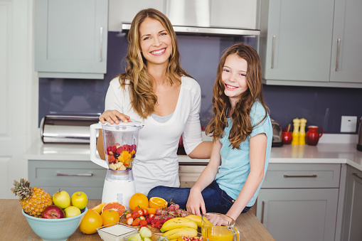 Portrait of happy mother and daughter preparing fruit juice at table in kitchen