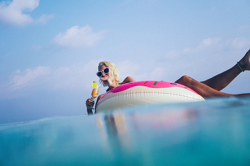 Attractive and beautiful woman is floating on float at the ocean and drinking cocktail. Float have shape of doughnut and it's pink colored. Cocktail is orange and have piece of lime on edge with straw in it. With one hand she is holding cocktail while with other is holding straw. Her short blonde hair is wet. She is wearing white sunglasses. Around her is crystal clear water and sky with some clouds. Calm environment is amazing for this hippy woman to get relaxed and refreshed.