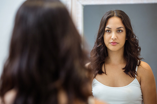 Beautiful young woman looking at herself in mirror at bathroom