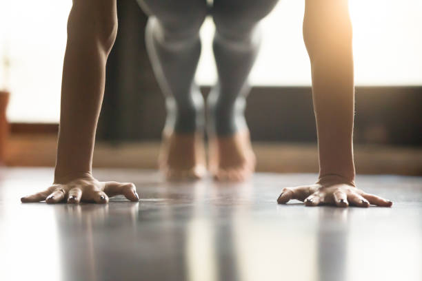 Young yogi woman standing in Plank pose, home interior backgroun Young woman practicing yoga, doing Push ups or press ups exercise, phalankasana Plank pose, working out, wearing sportswear, grey pants, indoor, home interior, living room floor. Close-up of hands yoga studio stock pictures, royalty-free photos & images