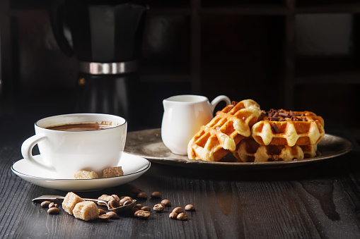 Homemade belgian waffles, white ceramic cup of coffee, milk, teaspoon and coffee beans. Dark rustic background. Space for text in the center