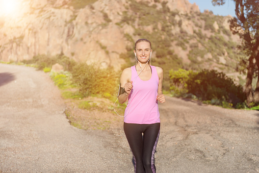 Horizontal color image of beautiful woman running in scenics rocky mountains and listening music. Vital young woman wearing pink tank top, arm band and wristwatch. Sportswoman enjoying beautiful sunset.