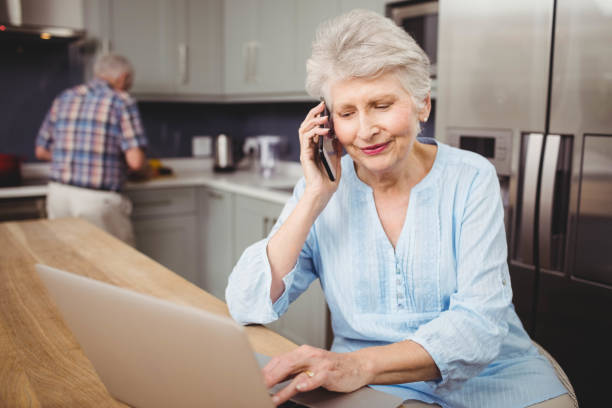 senior woman talking on phone while using laptop and man working in kitchen - cooking senior adult healthy lifestyle couple imagens e fotografias de stock