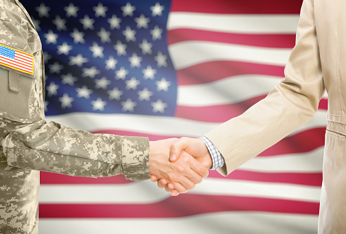 American soldier in uniform and civil man in suit shaking hands with national flag on background - United States