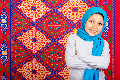 Ramadan Kareem - Happy Young Muslim girl celebrating Ramadan , wearing hijab and standing  in front of Islamic Arabian fabric traditional background