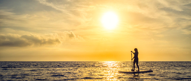 Side view of a woman paddle boarding on the sea with the sun in the background.