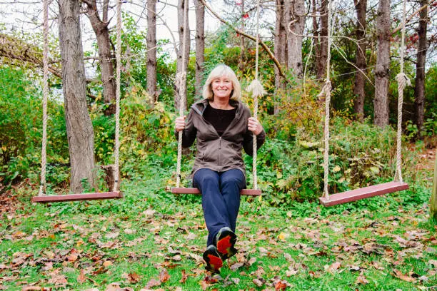 Senior woman having fun sitting and swinging on wooden handmade swing in the garden of her home in autumn. Active Senior, Real People, Lifestyle Portrait. Nova Scotia, Canada.