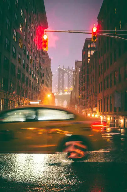 A yellow taxi drives by an intersection and gives a sense of motion to the image. Taken in DUMBO Brooklyn New York at night while it was raining. Manhattan Bridge can be seen in the background.