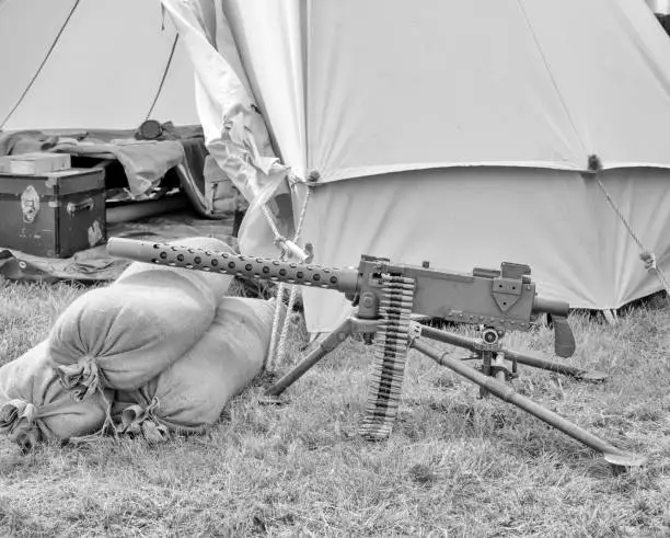 Old belt feed machine gun in front of a tent. Ground is grass. Sandbags in front. Machine gun is loaded. Low contrast black and white. Side view.