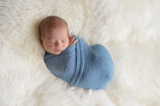 Sleeping, nine day old newborn baby boy swaddled in a light blue wrap. Shot in the studio on a white sheepskin rug.