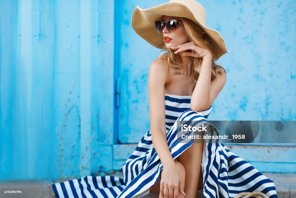 Summer portrait of a woman in a straw hat Woman portrait on blue background, sitting on the steps in a nice big straw hat and sun glasses, red lipstick and beautiful white teeth, with long blonde hair in a long striped dress with bare shoulders portrait of a model in the resort Summer Stock Photo
