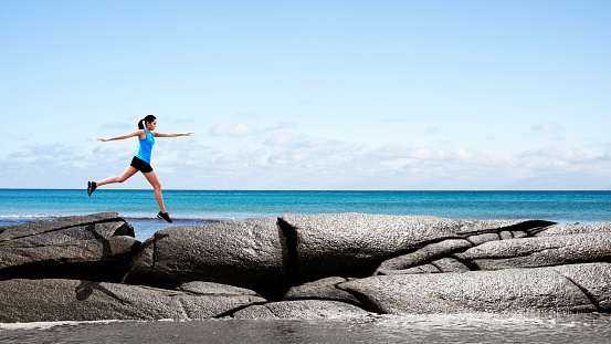 Woman in running clothes jumping rock to rock by the ocean with waves splashing up against the rocks. Location: Nova Scotia