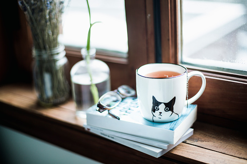 A pretty cup of tea on which there is a  cat image on a stack of books with glasses and flowers beside