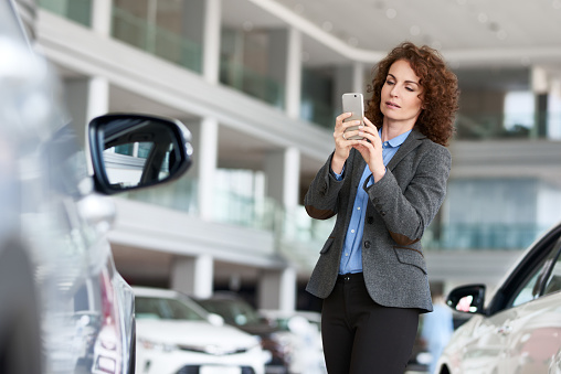 Concentrated mid adult woman using smartphone to take photo of car which she wants to buy