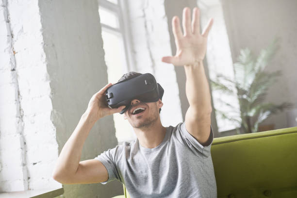 istock Hansome european male experiencing virtual reality using VR headset glasses, sitting on couch at home and throwing hand in air, gesturing. Young  man wearing oculus or goggles playing video games 675823810