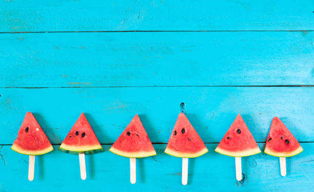 A red, ripe slice of watermelon on picnic table outdoors in summer season. A picnic basket is behind the watermelon, which lays on a red checked tablecloth. Beautiful and sunny park, nature scene in the background.  No people in image.  Memorial Day, July 4th, or Labor Day family picnic concept. Copyspace to right. 