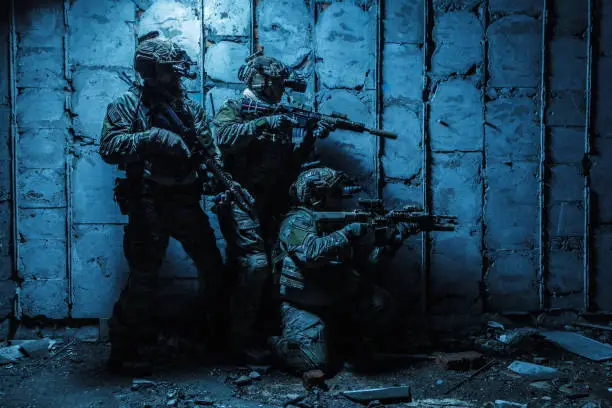 Squad of Army Rangers with rifle and machine gun moving along the concrete wall of ruined destroyed building on mission. They are ready to start firing if enemy appear. Outdoor location shot, darkness of night, dim light