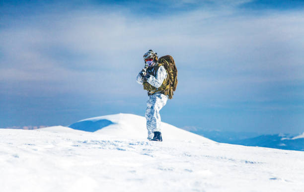 Army serviceman in winter camo somewhere in the Arctic. He wears chest rig and huge backpack, suffers from the extreme cold and strong wind, but endures while mission continues. Blue sky background