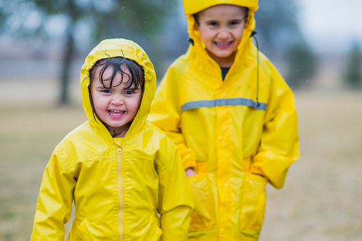 A young Caucasian brother and sister are outdoors in a park. It is raining. They are wearing bright yellow raincoats. They are both standing and smiling.