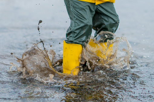 A young Caucasian boy is outdoors in a park. It is raining. He is wearing a bright yellow raincoat. He has bright yellow rubber boots. He is splashing in a puddle.