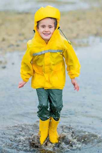 A young Caucasian boy is outdoors in a park. It is raining. He is wearing a bright yellow raincoat. He has bright yellow rubber boots. He is splashing in a puddle and smiling at the camera.