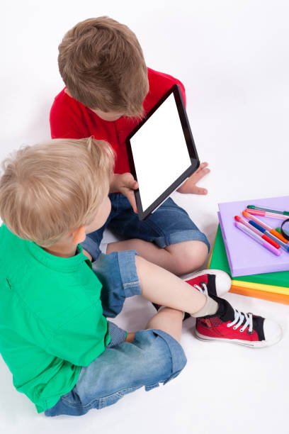 istock Two schoolchildren sitting on a white background, surrounded by books and looking at tablet. 675947968