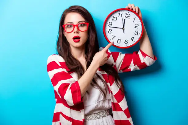 Photo of beautiful surprised young woman with clock standing in front of wonderful blue background