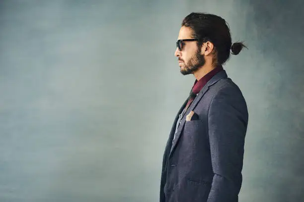 Studio profile shot of a stylishly dressed handsome young man with a ponytail wearing sunglasses