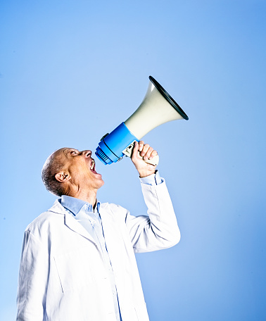 A mature, balding, professional man wearing a white, seen in profile, looks up into blue copy space over the bullhorn he's shouting into.