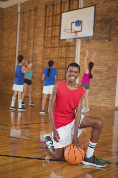 istock Smiling school boy kneeling with a basketball while team playing in background 676023890