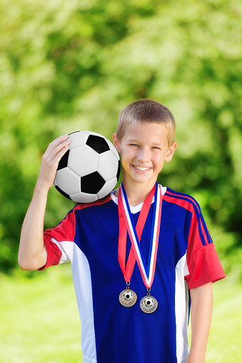 Portrait of a Caucasian teen boy in soccer uniform, holding up a soccer ball on a grass lawn.