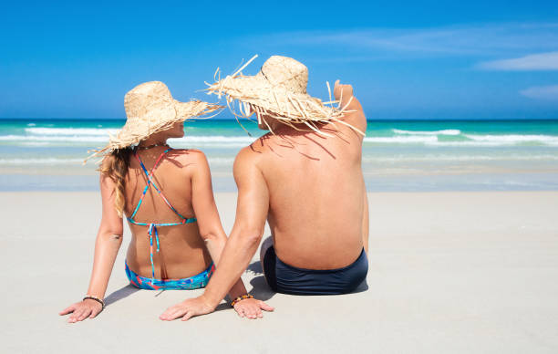 istock Cheerful couple in straw hats, talking while sitting on white sand looking at ocean 676222608