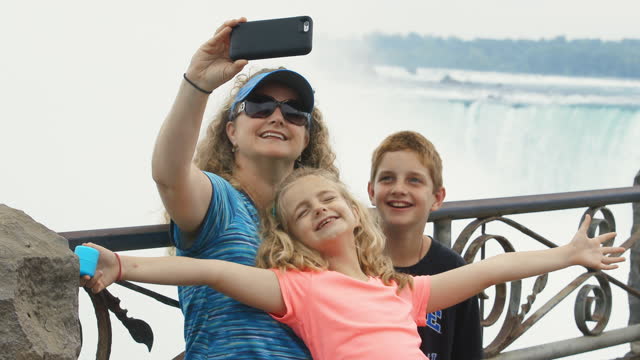 MS. Mom takes smartphone selfies with smiling kids overlooking scenic Niagara Falls.