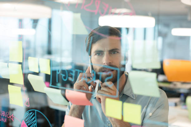 istock Businessman using mobile phone while looking at plans written on glass 676234548