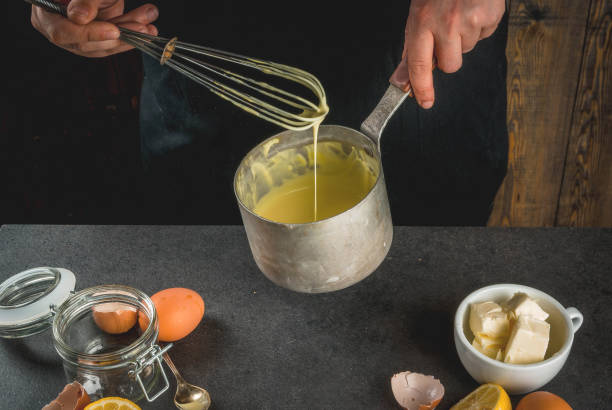Basic Hollandaise sauce Traditional basic sauces. French cuisine. Person in the frame is preparing Hollandaise sauce. In a metal saucepan, with ingredients - eggs, butter, lemons. On black stone table. Female hands. hollandaise sauce stock pictures, royalty-free photos & images