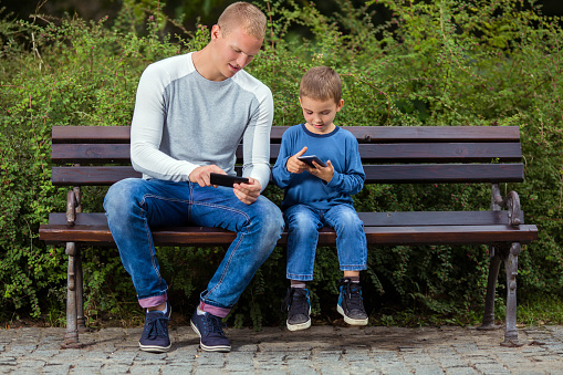 Young father and his cute little son are playing video games while relaxing on a park bench