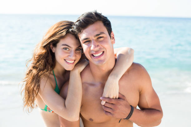 Happy young couple standing on shore at beach Close-up portrait of happy young couple standing on shore at beach male swimsuit standing arm around stock pictures, royalty-free photos & images