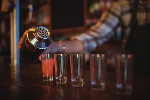 Waiter pouring cocktail drink into shot glasses at counter in bar