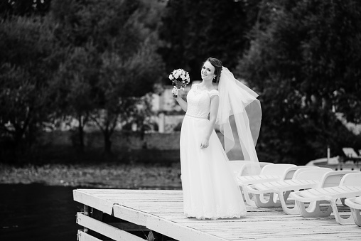 Portrait of cute brunette bride with wedding bouquet at hands on pier. Black and white photo.